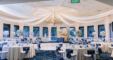 An elegant reception setup with round tables, floral centerpieces and draped fabric under a sparkling chandelier