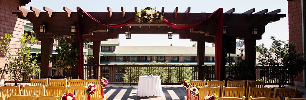 Rows of wooden chairs flank a petal covered aisle leading up to a pergola decorated with flowers and a floor length fabric swag