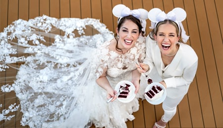 Two smiling brides wearing Minnie Mouse ear headbands and eating red velvet cake