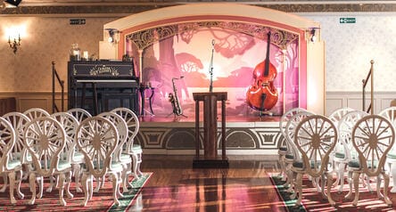 A room with rows of chairs set in front of a podium features a stage with a piano and jazz instruments on stands