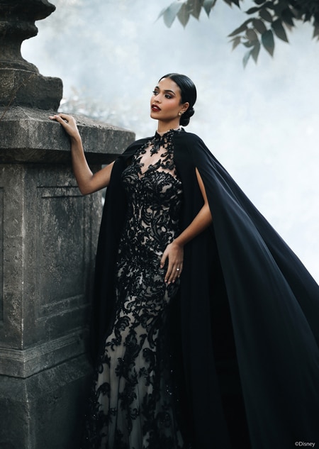 A woman posing by a stone structure wearing a fitted gown with intricate lace details and a flowing cape