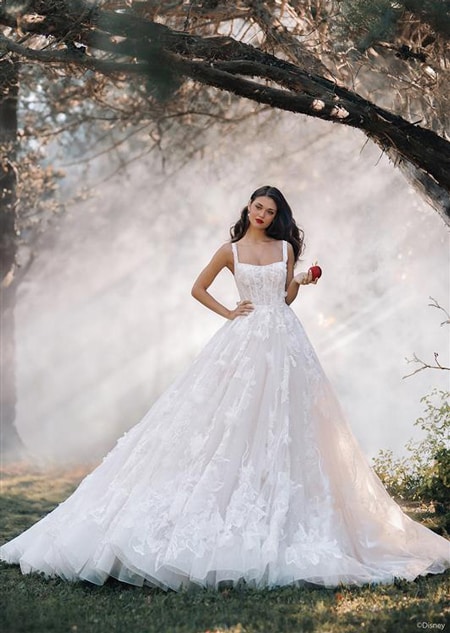 A woman posing in a forest wearing a fairy tale wedding gown with floral details holding an apple 