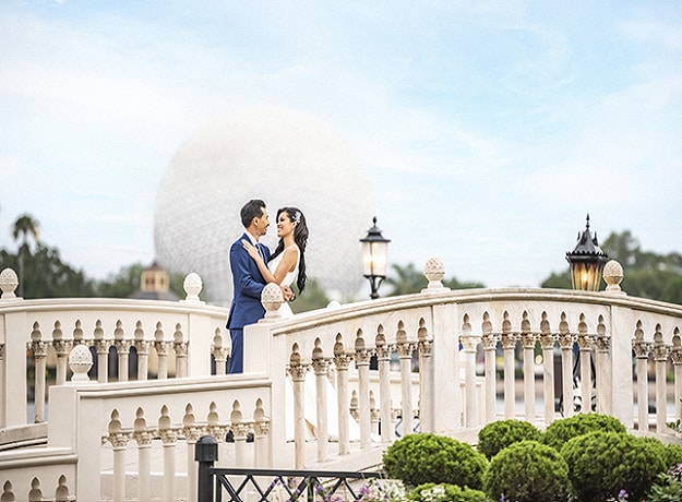 A couple in wedding attire embrace on an ornate bridge with Spaceship Earth in the background 
