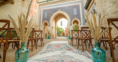 An outdoor Moroccan themed ceremony setup with a rug lined aisle, wooden chairs and decorative vases near an ornate archway