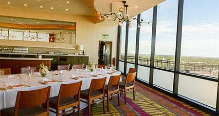 An indoor dining setup with a long table, wooden chairs and floor to ceiling windows overlooking scenic views