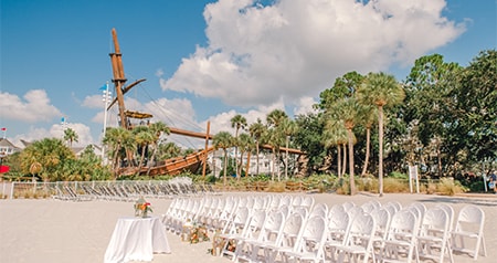 A beachfront ceremony setup with chairs, a decorated table and a shipwreck themed backdrop surrounded by palm trees