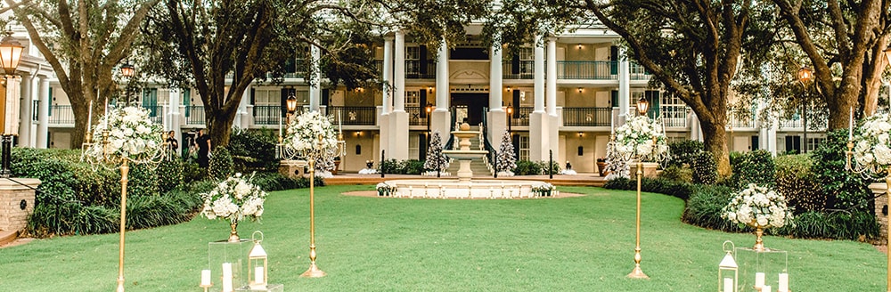 A stately manor with oak trees and a fountain is set up for an outdoor wedding on the lawn