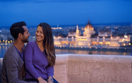 A man and a woman cuddle on a balcony at dusk with the city in the background