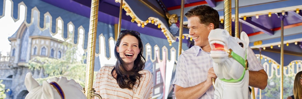 A woman and a man smile widely as they sit atop wooden horses on an old fashioned carousel ride
