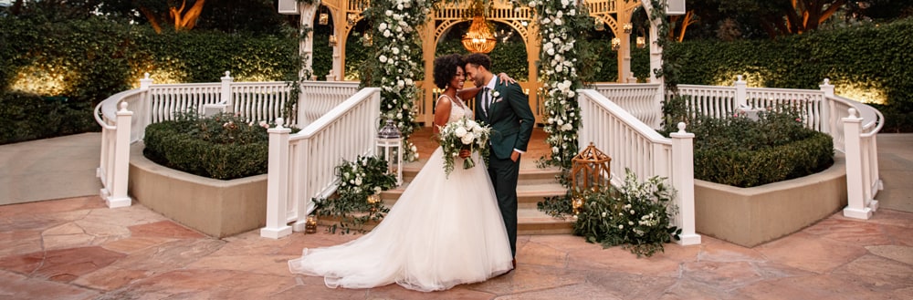 A bride and groom embracing in front of an elegant gazebo decorated with chandeliers and florals 