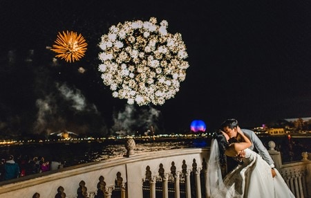 Couple kissing on balcony with Epcot's Spaceship Earth and fireworks in the background.
