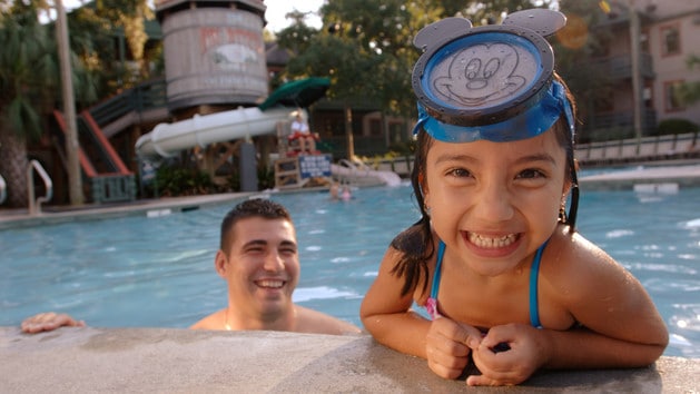 A little girl with a Mickey snorkel mask leans on the side of the pool while her dad watches