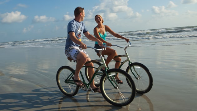 A man and woman ride bikes on the wet sand of the beach as small waves break