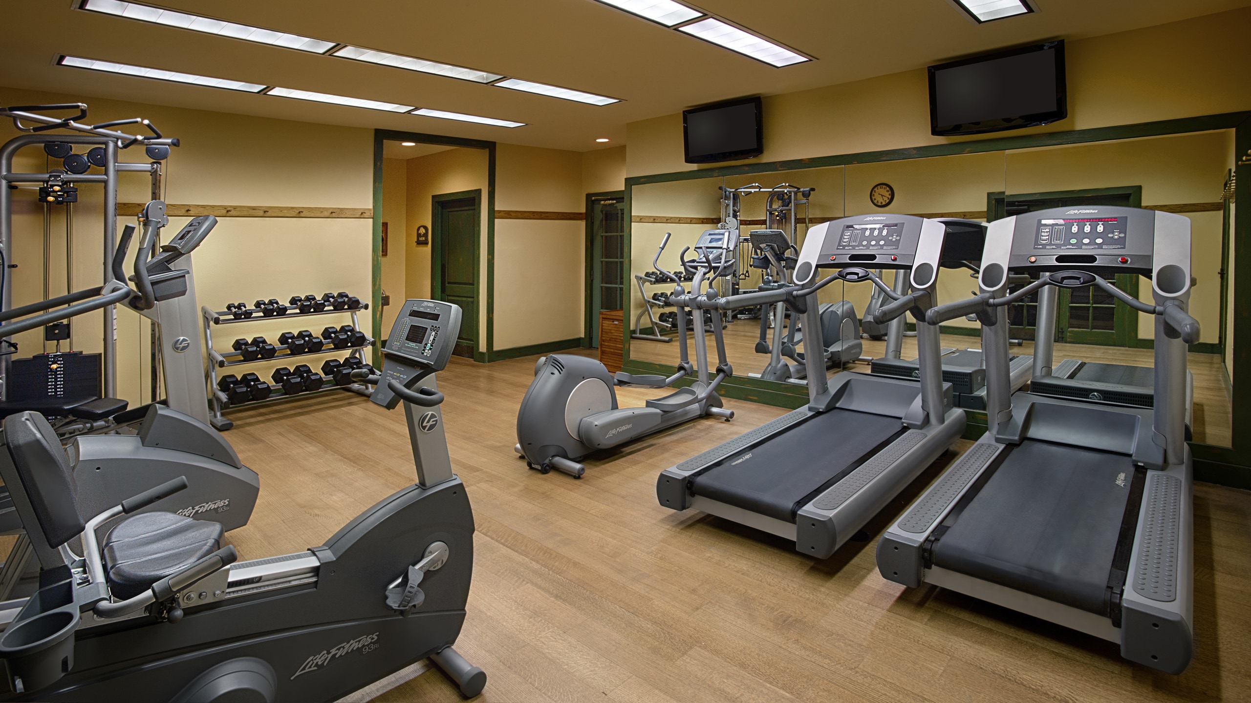 A mirrored gym area with treadmills, ellipticals, a workout bench and a TV mounted on the wall