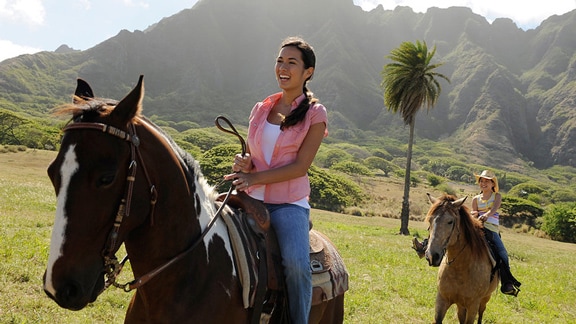 Two women riding horses across a field at the base of a mountain