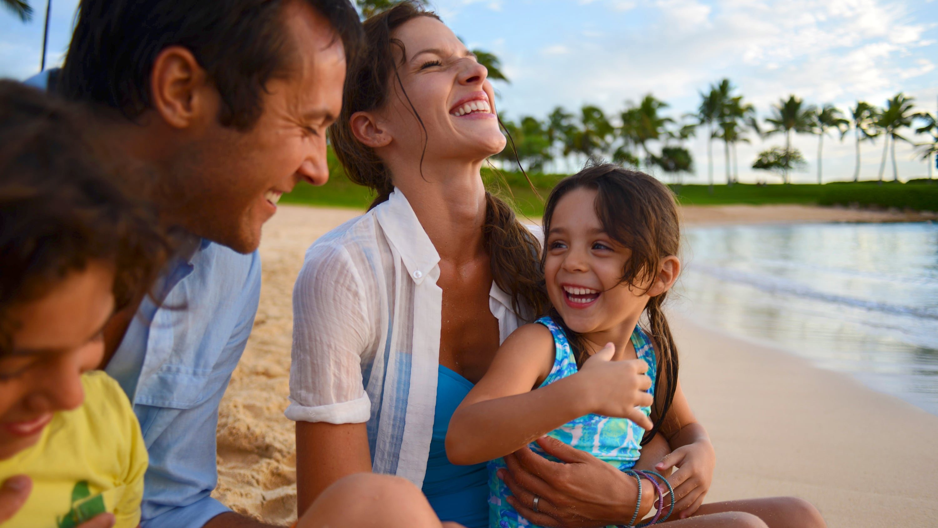 A father and mother hold their 2 children as they sit on the shore of a beach near calm waves