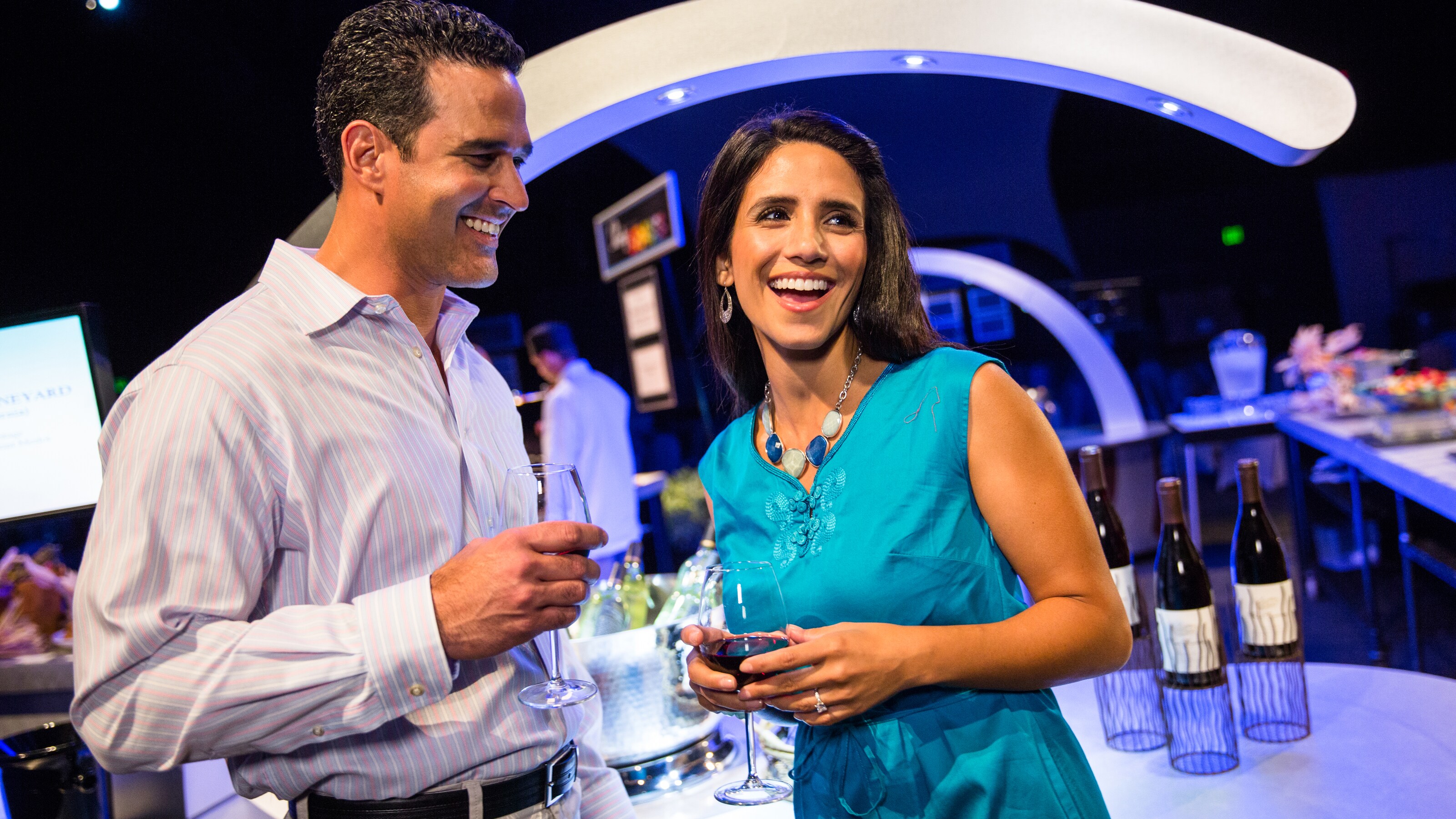 A man and woman laughing while holding glasses of wine with food and wine in the background