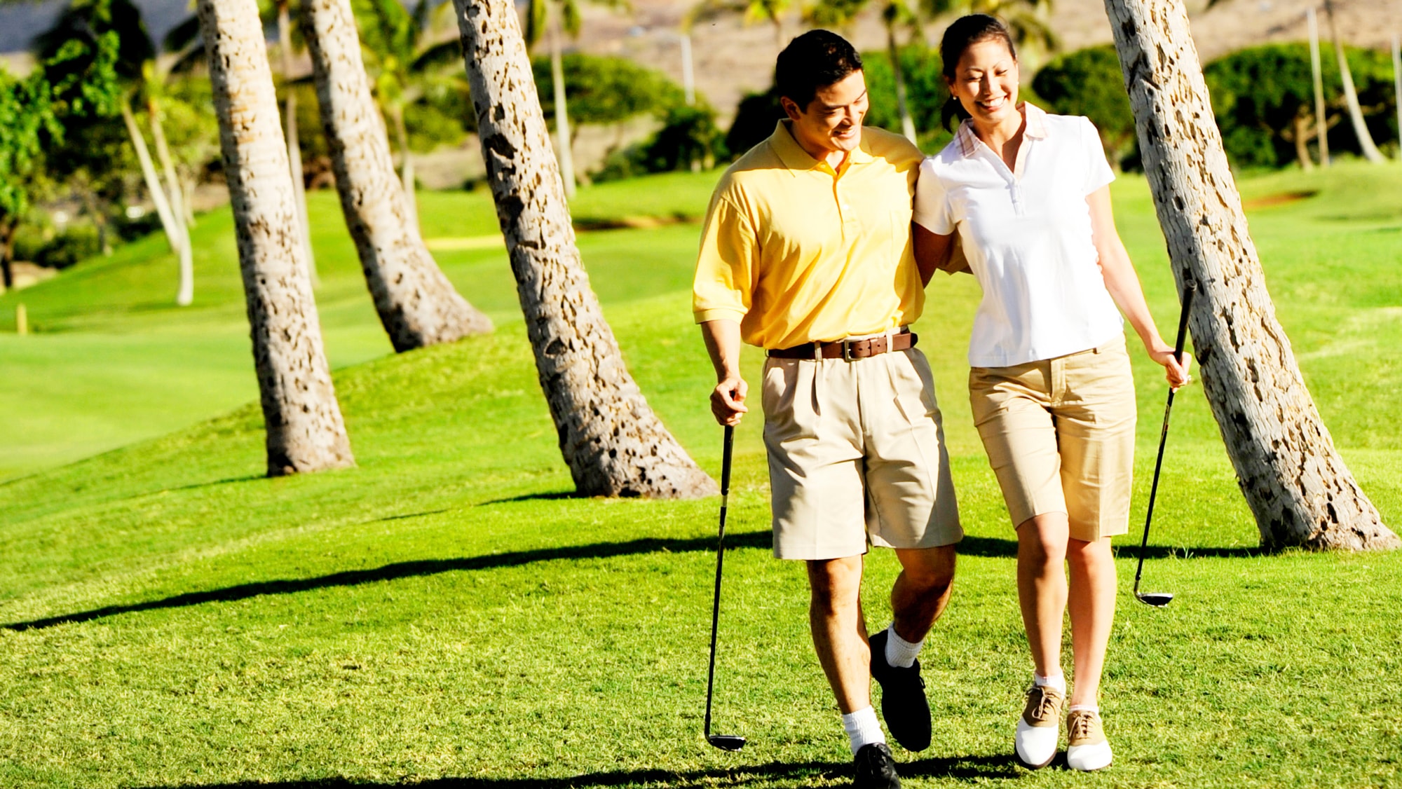 A man and a woman with their arms around one another carry golf clubs as they walk down a fairway