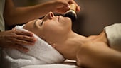A woman lying face-up on a spa table while a spa therapist applies a brush to her face