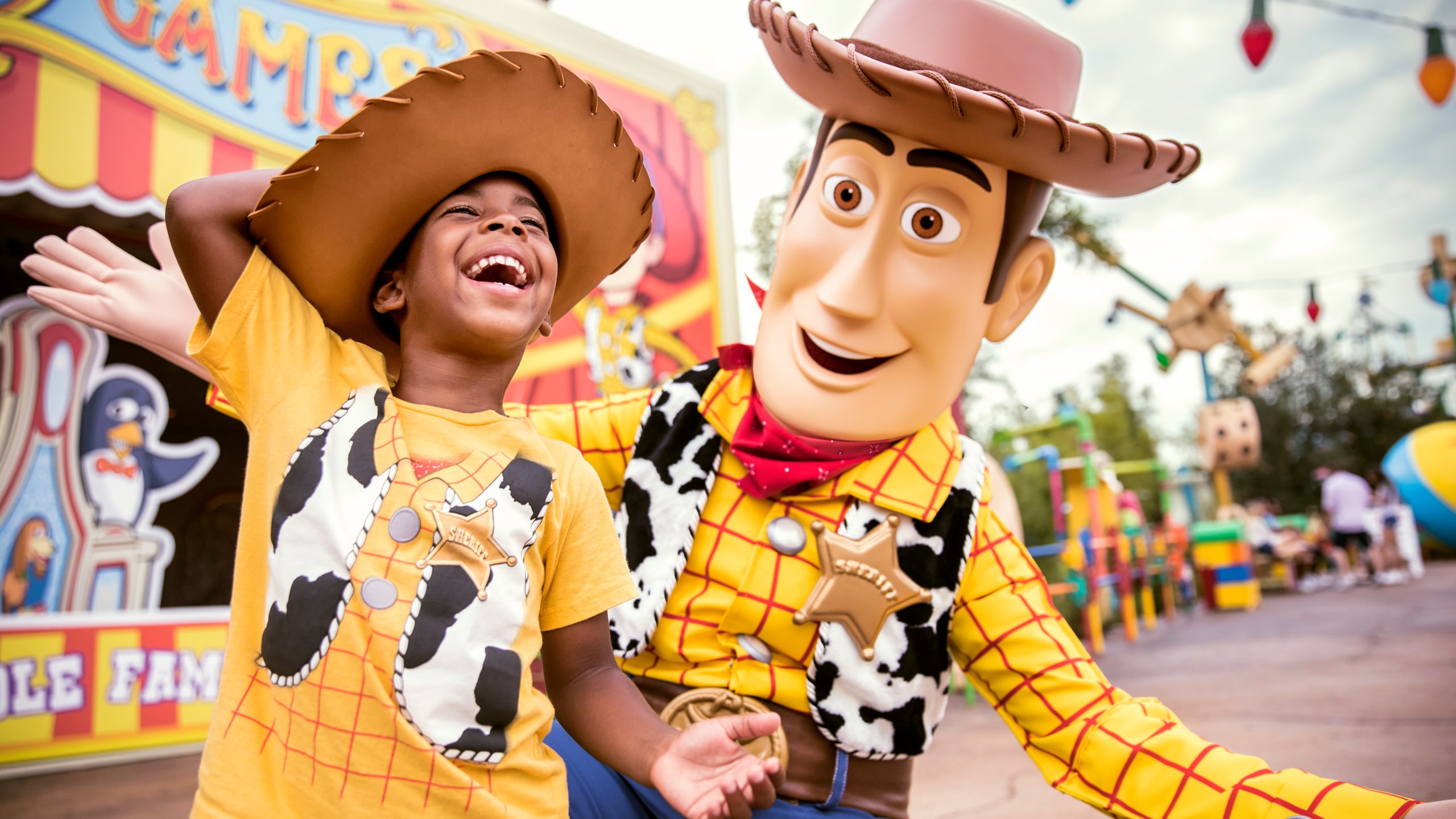 A young boy wearing a Woody themed tee shirt and cowboy hat laughs as a interacts with Woody inside of Toy Story Land