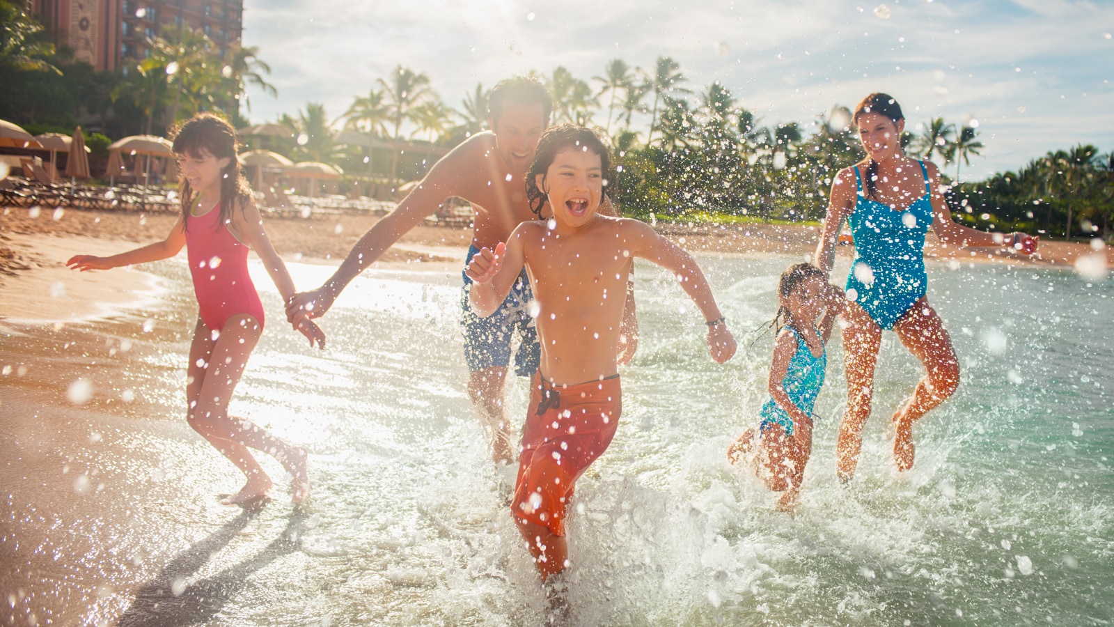 A mother, father and 3 children splash along the ocean shoreline on a sunny day