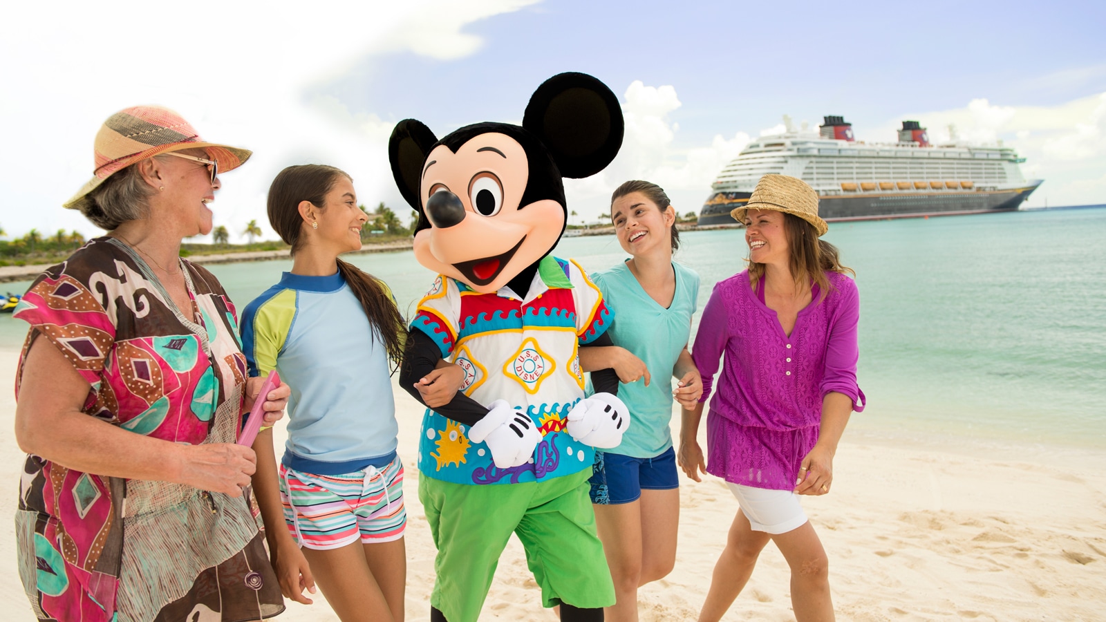 Mickey Mouse walks with 4 women on a beach with a Disney Cruise Line ship in the background