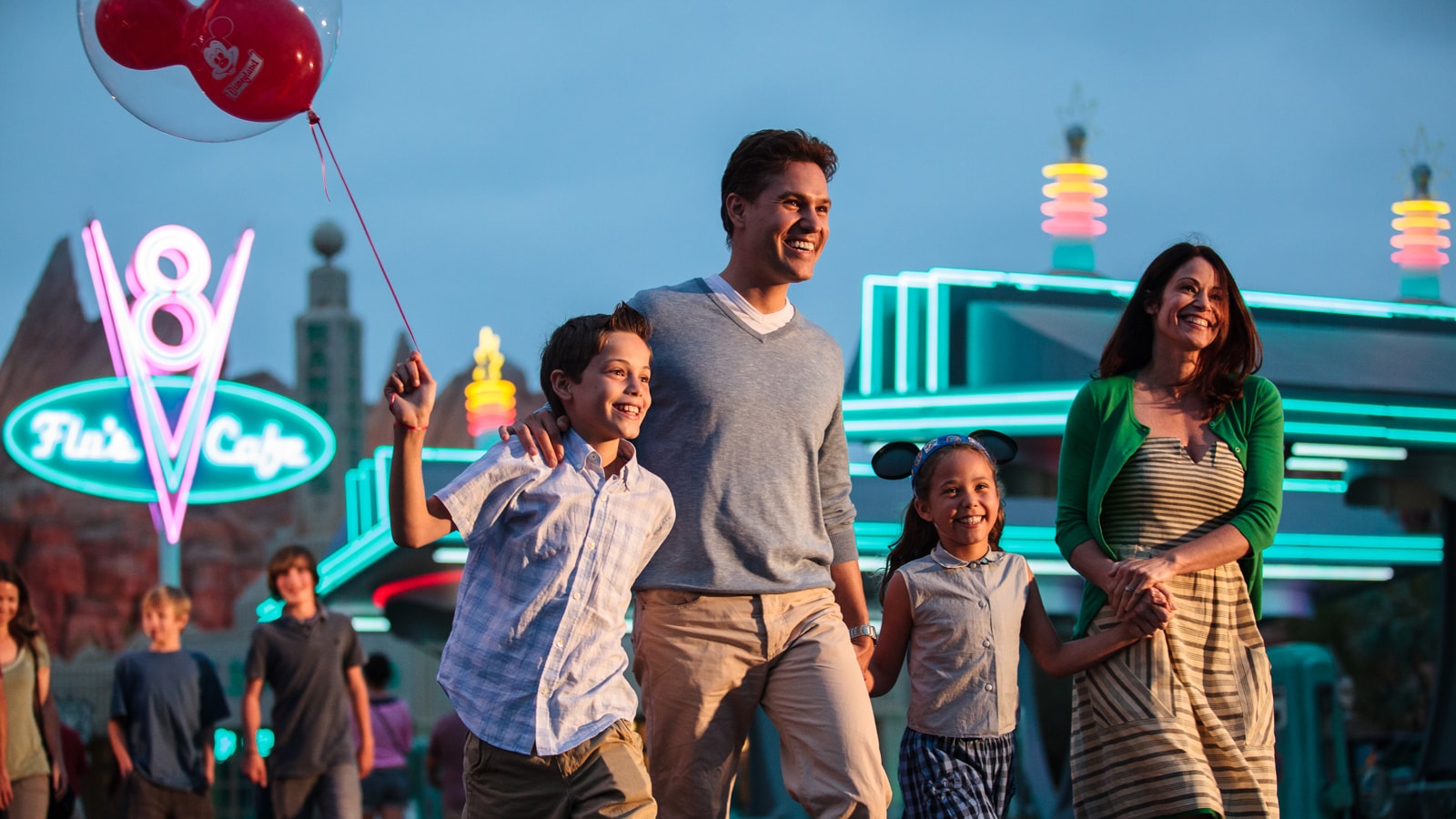 A family of 4 walks in front of building with neon sign that says 'Flo's Cafe' at dusk