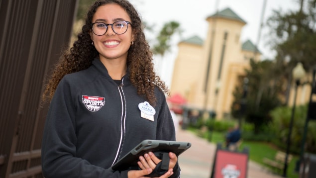 A Cast Member smiling and holding a tablet at the ESPN Wide World of Sports Complex main entrance 