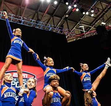 Eight girls from the Wolves cheer team in a formation where 5 girls hold 3 girls on their shoulders in front of an ESPN Wide World of Sports banner