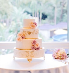 A 3 tiered ornate wedding cake with a ′Tangled′ design on a cake stand near a bouquet of roses