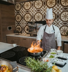 A chef in an apron and hat in an upscale kitchen space sautéing vegetables in a pan with a flame