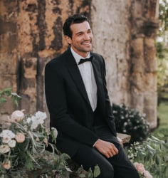 A man in a black tuxedo and bow tie smiling while sitting near floral arrangements  
